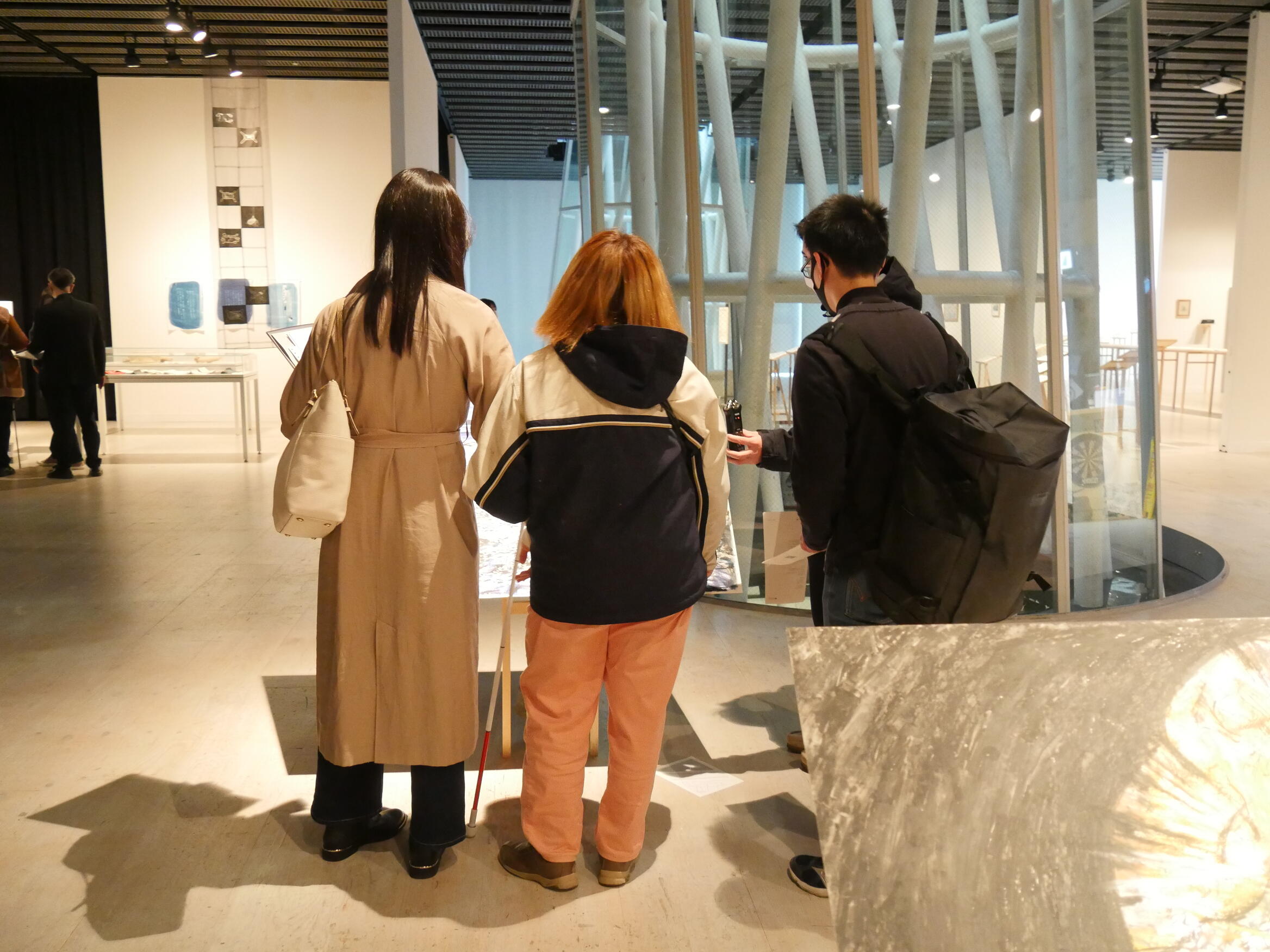 菊池聡太朗さんの作品を鑑賞している様子。白杖を持った見えない学生一人と見える学生3人が荒れ地を描いたドローイングの前に立って会話をしている